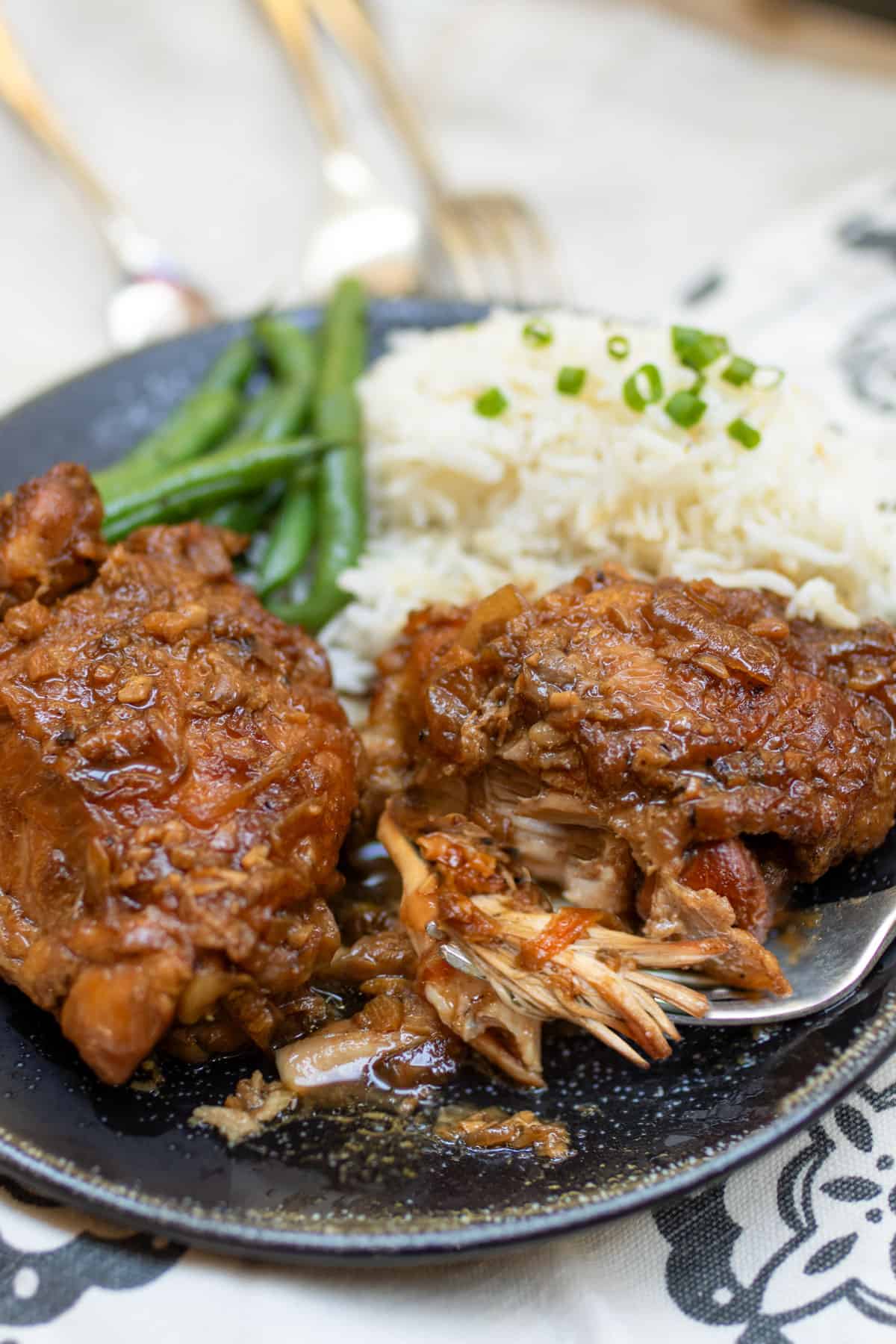 Two pieces of Filipino chicken adobo, braised chicken thighs, covered in rich thick adobo sauce, with rice and green beans on the side. A fork is pulling some tender looking chicken from a thigh.