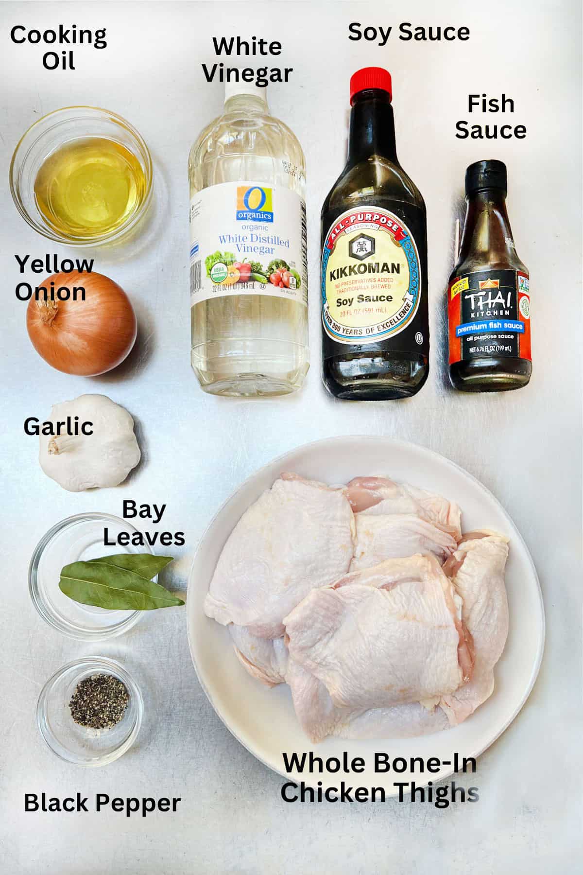 ingredients with labels on a counter: bowl of cooking oil, bottles of white vinegar, soy sauce and fish sauce, 1 yellow onion, 1 whole garlic bulb, 2 bay leaves, small bowl of black pepper, and a plate of raw bone-in chicken thighs.