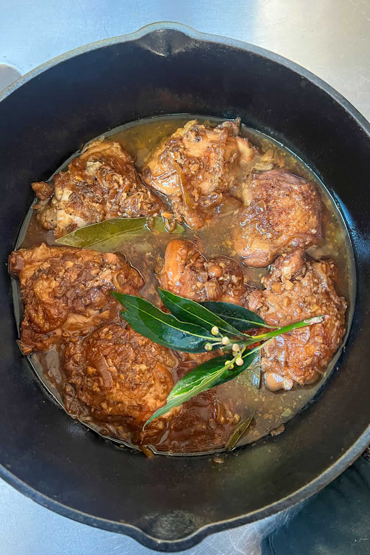 7 braised chicken thighs in a cast iron dutch oven, bathed in rich brown adobo sauce with sprig of bay leaves to garnish.