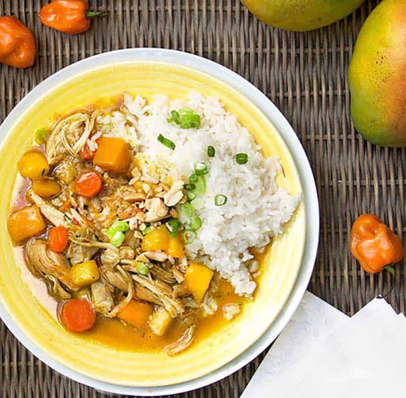 colorful Jamaican curry chicken stew with carrots, butternut squash and mango, in a white bowl with small bowls on the side of chopped peanuts and chopped scallions 