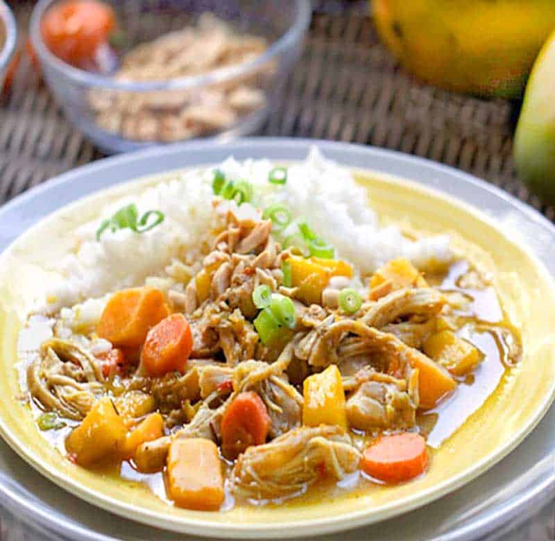 colorful Jamaican curry chicken stew with carrots, butternut squash and mango, in a white bowl with small bowls on the side of chopped peanuts and chopped scallions 