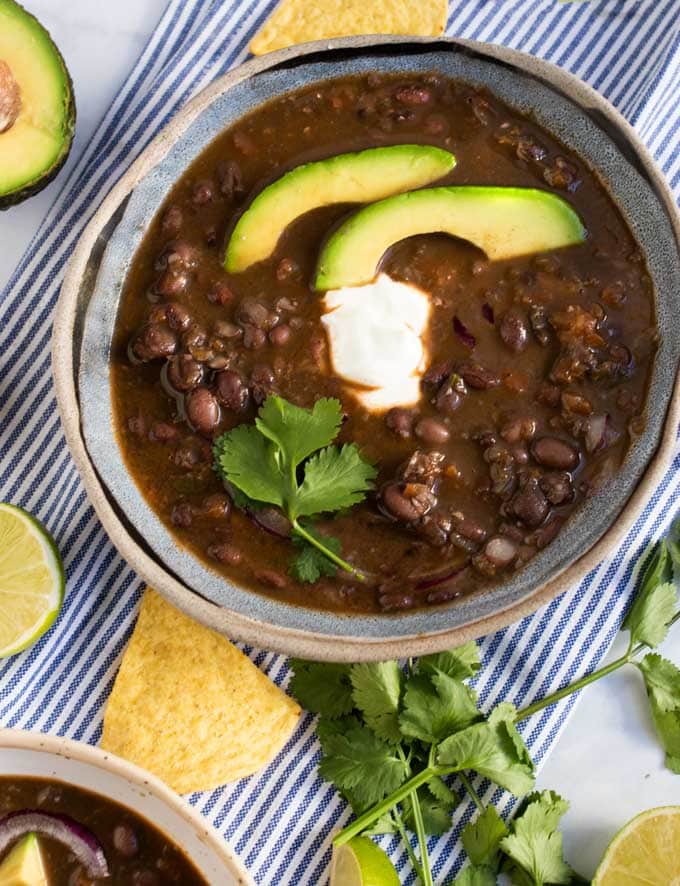 A striped dish towel under a blue bowl filled with spicy black bean soup garnished with two avocado slices, sour cream and a sprig of cilantro. 