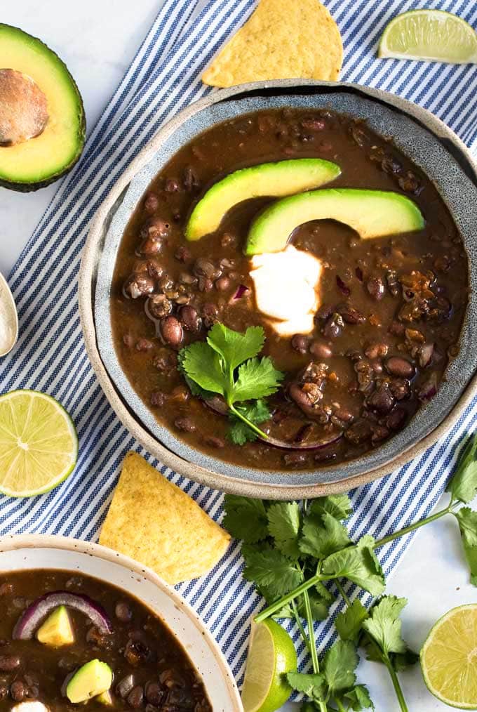 A striped dish towel under a blue bowl filled with spicy black bean soup garnished with two avocado slices, sour cream and a sprig of cilantro. 
