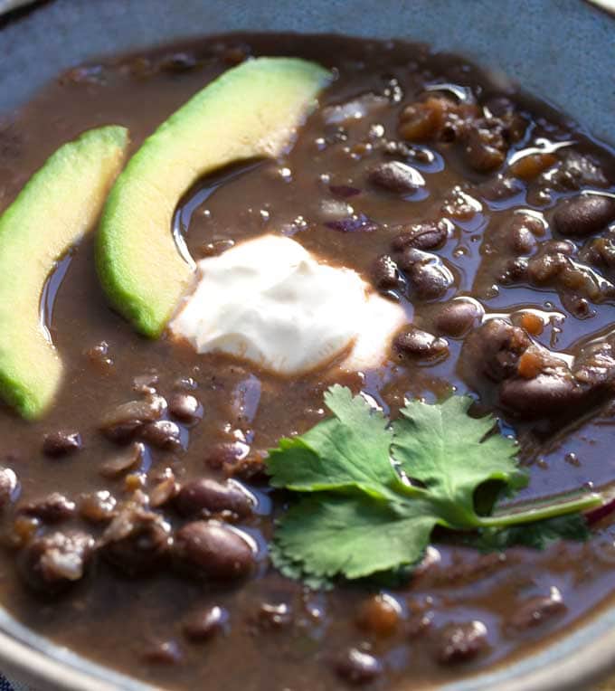 close up of a blue bowl filled with spicy black bean soup garnished with two slices of avocado, a sprig of cilantro and a dollop of sour cream