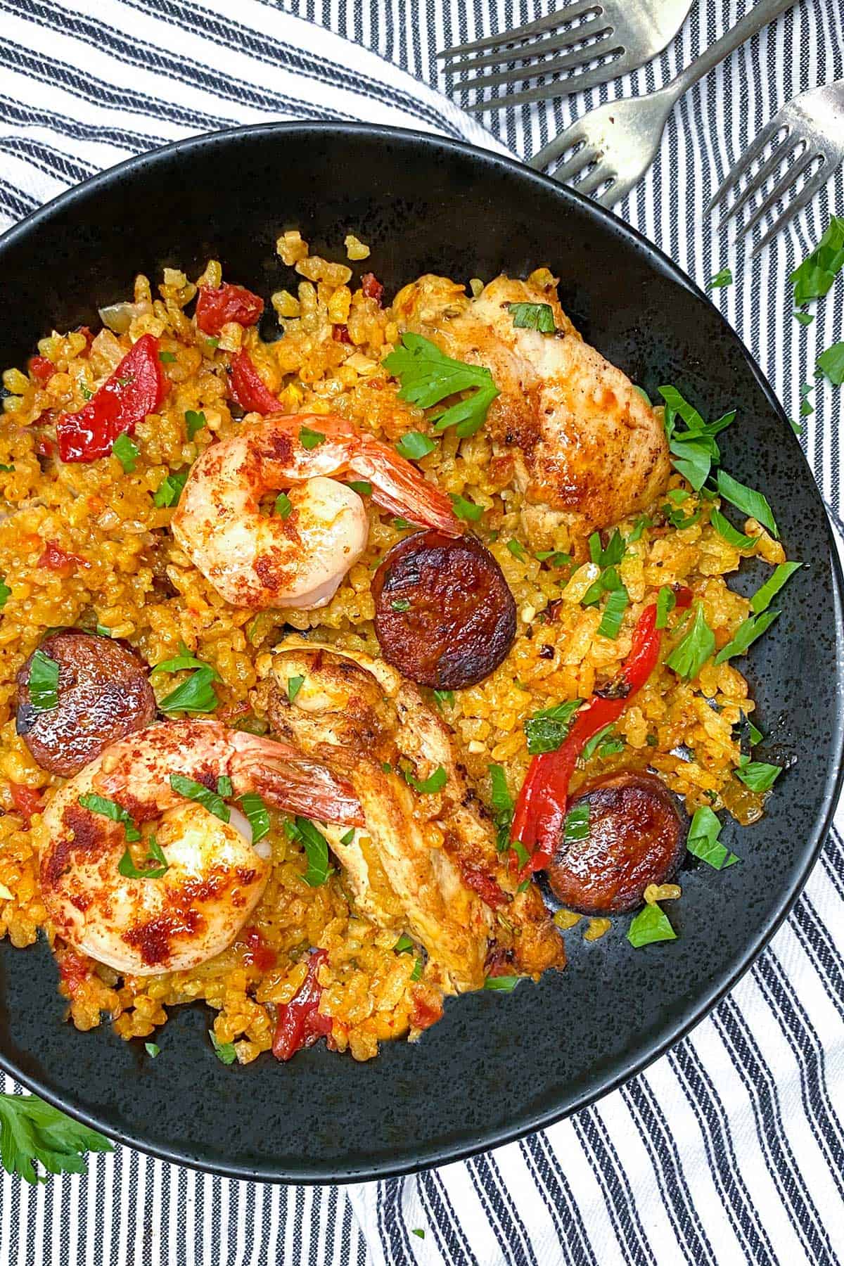 overhead looking into a black bowl filled with Spanish paella with yellow rice, a few shrimp, some chicken strips, 3 sliced of sausage and parsley sprinkled on top