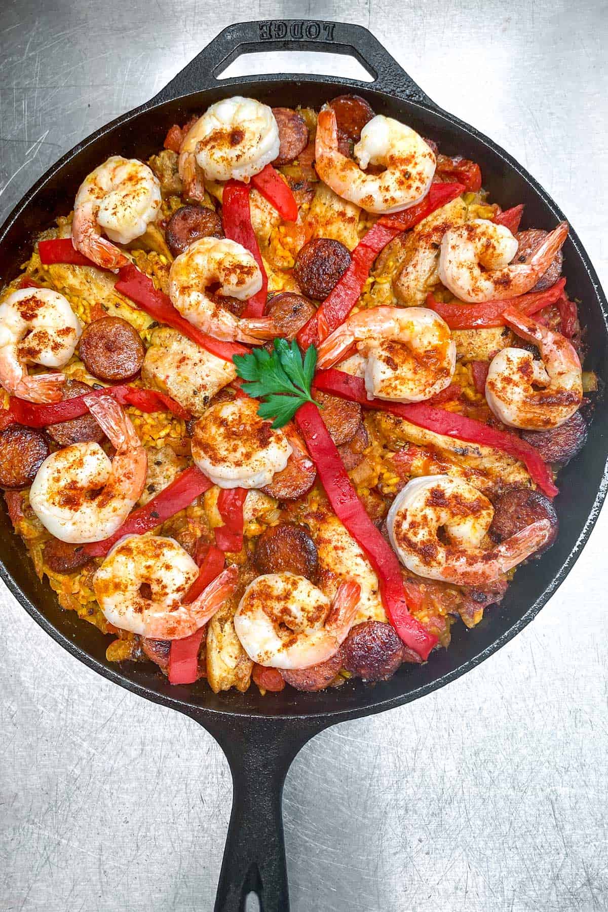 shrimp, chicken and sausage paella in a cast iron pan topped with strips of roasted red pepper and a sprig of parsley in the middle