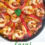 Pinterest pin with overhead shot of a cast iron skillet filled with paella: shrimp, sausages, chicken, strips of pimiento and a parsley leaf in the middle