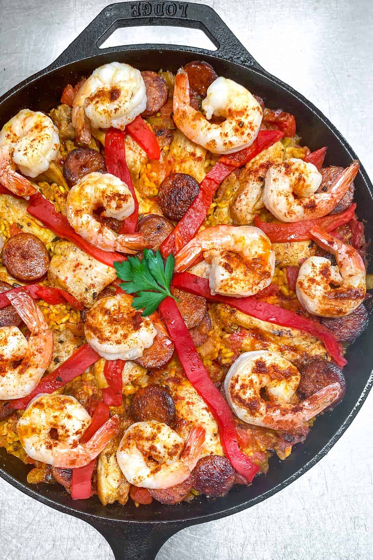 overhead shot of a cast iron skillet filled with paella: shrimp, sausages, chicken, strips of pimiento and a parsley leaf in the middle