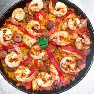overhead shot of a cast iron skillet filled with paella: shrimp, sausages, chicken, strips of pimiento and a parsley leaf in the middle