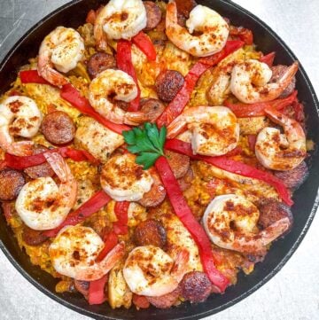 overhead shot of a cast iron skillet filled with paella: shrimp, sausages, chicken, strips of pimiento and a parsley leaf in the middle