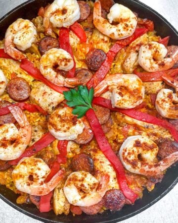 overhead shot of a cast iron skillet filled with paella: shrimp, sausages, chicken, strips of pimiento and a parsley leaf in the middle