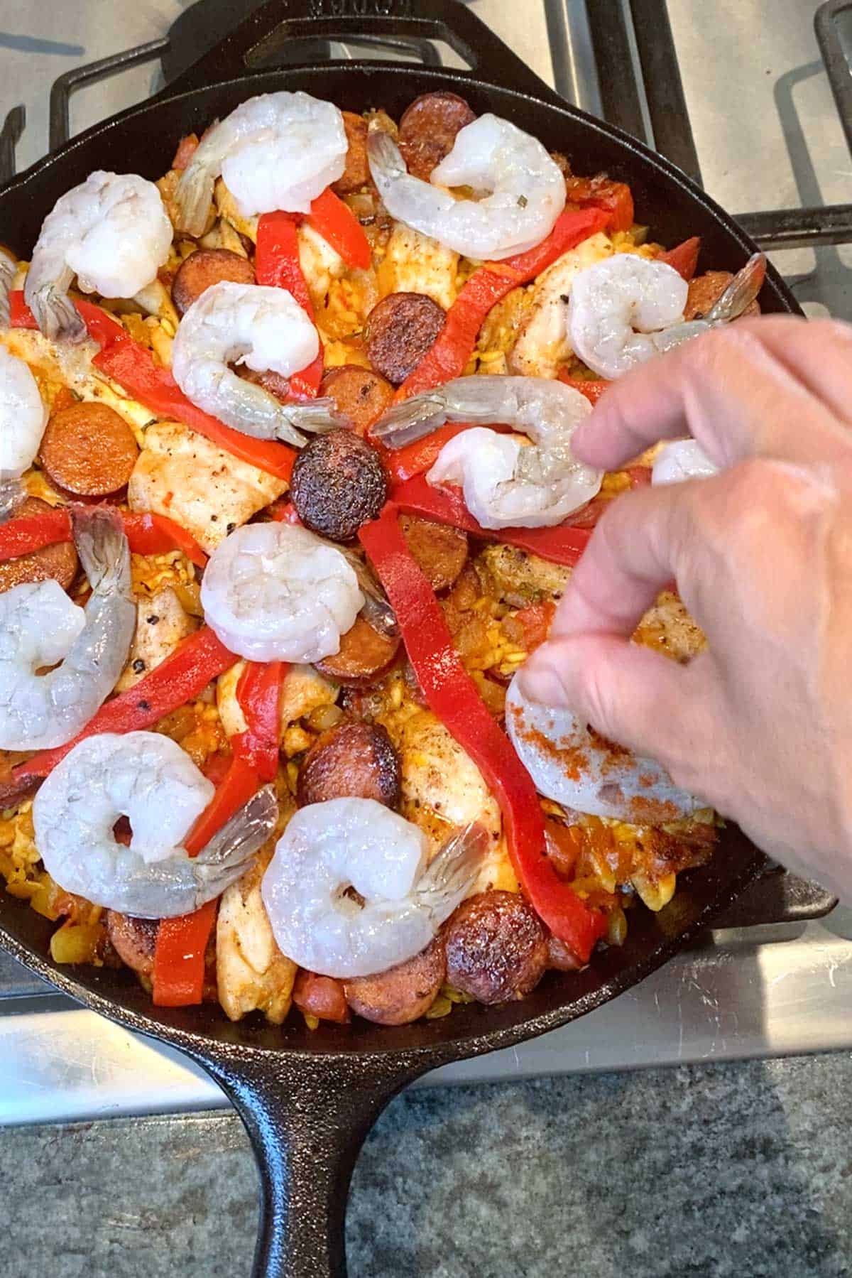 Overhead shot of a black cast iron skillet with raw shrimp on top of saffron rice and a hand sprinkling paprika on the shrimp
