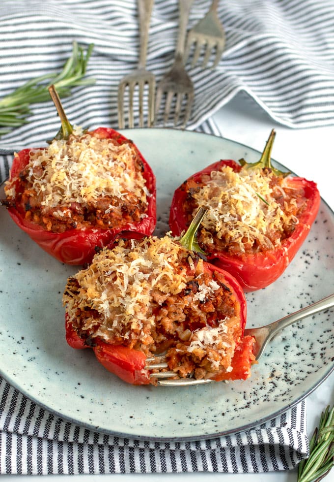 three pork and parmesan stuffed red peppers on a plate with three forks in the background and a sprig of rosemary and a striped dish towel