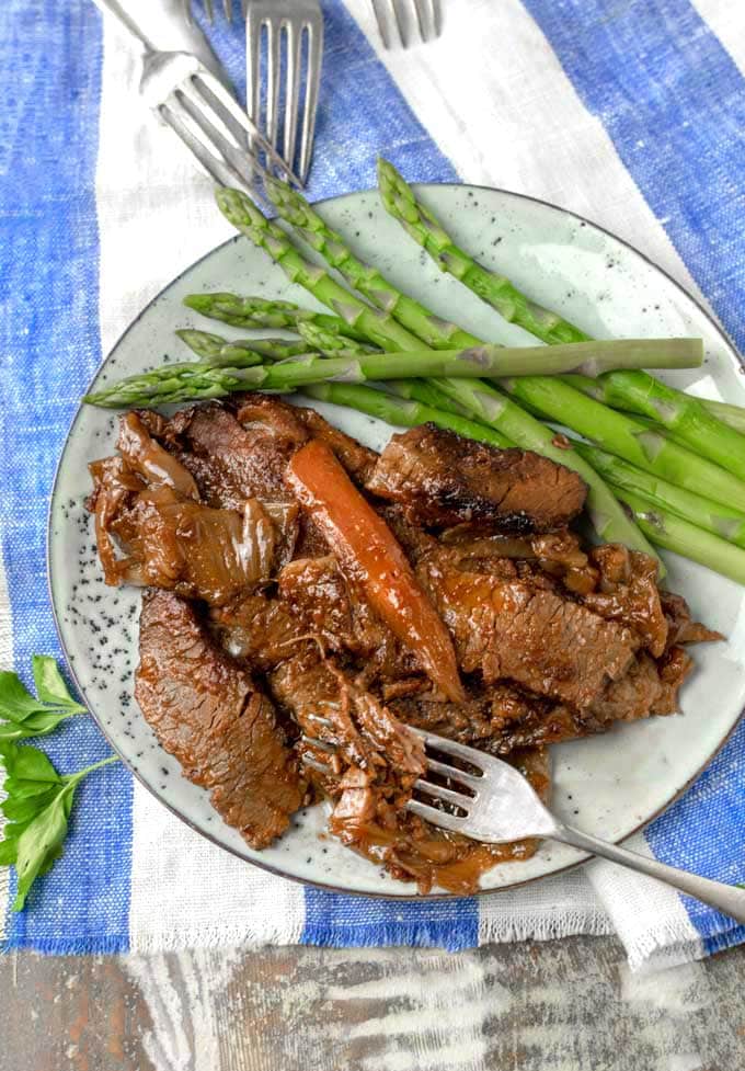 A plate of Nach Waxman's brisket with a carrot on top and asparagus on the side, with a blue and white striped cloth napkin and four forks