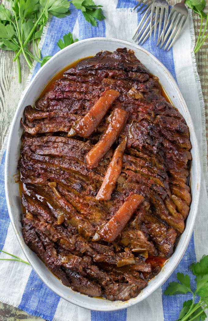 A serving dish with Nach Waxman's Brisket, cooked, sliced and topped with four cooked carrot pieces, on a blue and white striped napkin garnished with parsley and forks