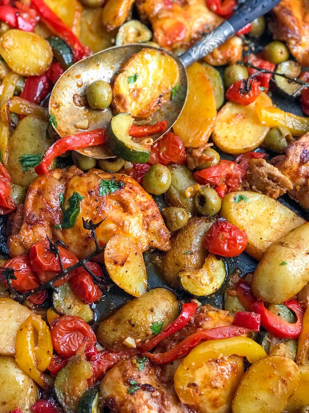 close up of a baking dish with baked boneless chicken thighs, sliced baby potatoes, red and yellow bell peppers, chunks of zucchini, green olives and cherry tomatoes. 