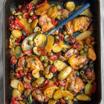 rimmed baking sheet topped with baked chicken thighs, potatoes, red bell peppers, zucchini and green olives.
