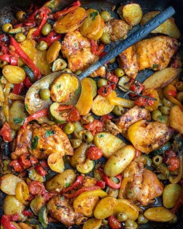 rimmed baking sheet topped with baked chicken thighs, potatoes, red bell peppers, zucchini and green olives.