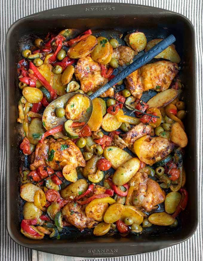 A black baking dish with baked boneless chicken thighs, sliced baby potatoes, red and yellow bell peppers, chunks of zucchini, green olives and cherry tomatoes.