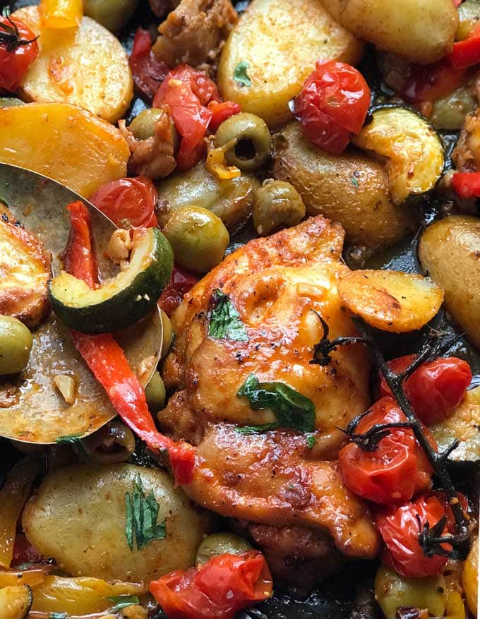 close up of a baked chicken thigh with sliced potatoes, bell peppers, cherry tomatoes and green olives