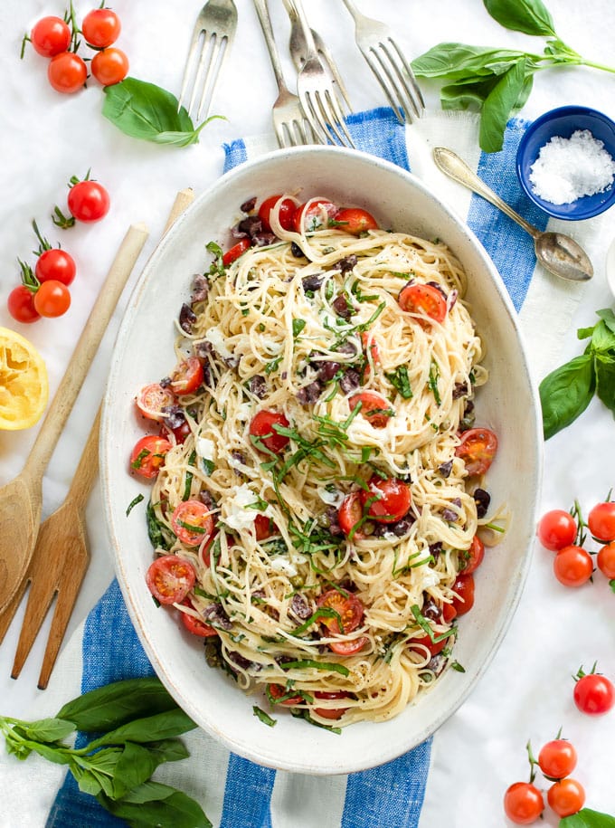 an oval bowl filled with angel hair pasta with cherry tomato and goat cheese no-cook sauce, on a blue and white striped dish towel scatterd with basil leaves and clusters of cherry tomatoes