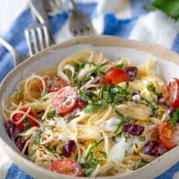 bowl of angel hair pasta with cherry tomatoes, goat cheese, basil and olives, on a blue and white striped dish towel with four forks pointing to the bowl and lemons in the background