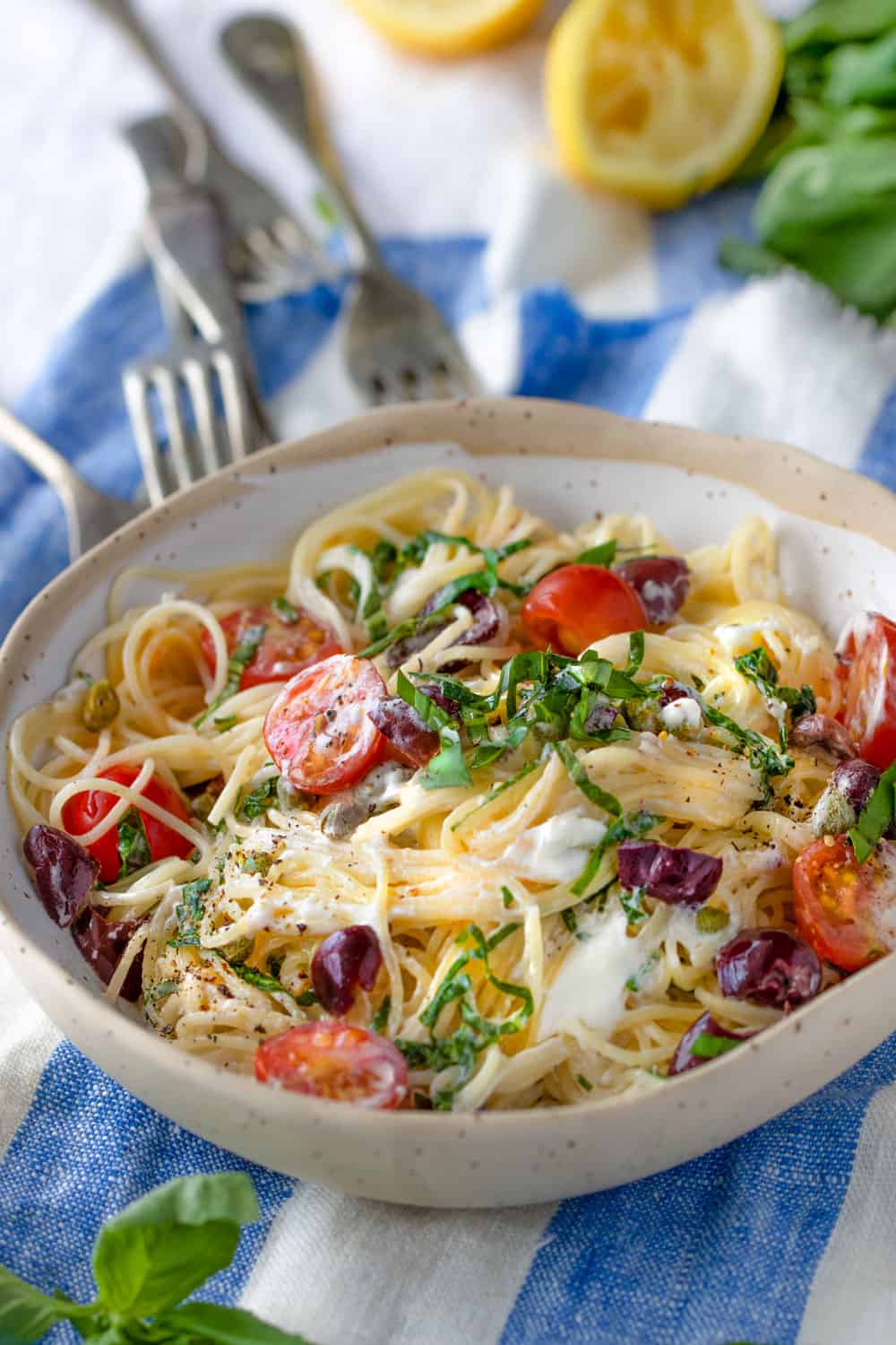 bowl of angel hair pasta with cherry tomatoes, goat cheese, basil and olives, on a blue and white striped dish towel with four forks pointing to the bowl and lemons in the background