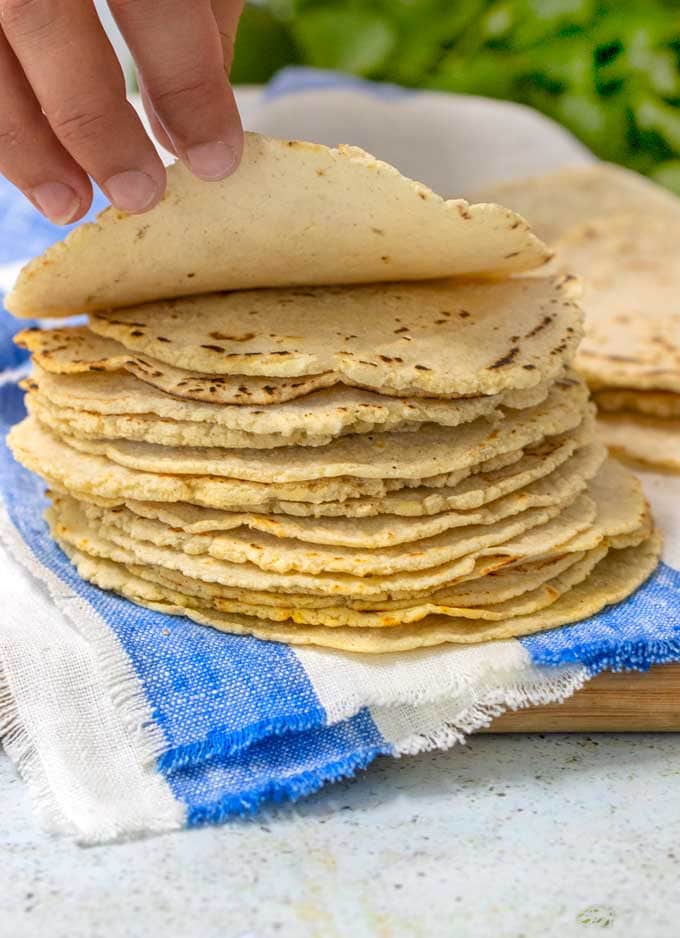 A stack of homemade corn tortillas on a blue and white striped dish towel, with a hand lifting up the edge of the top tortilla