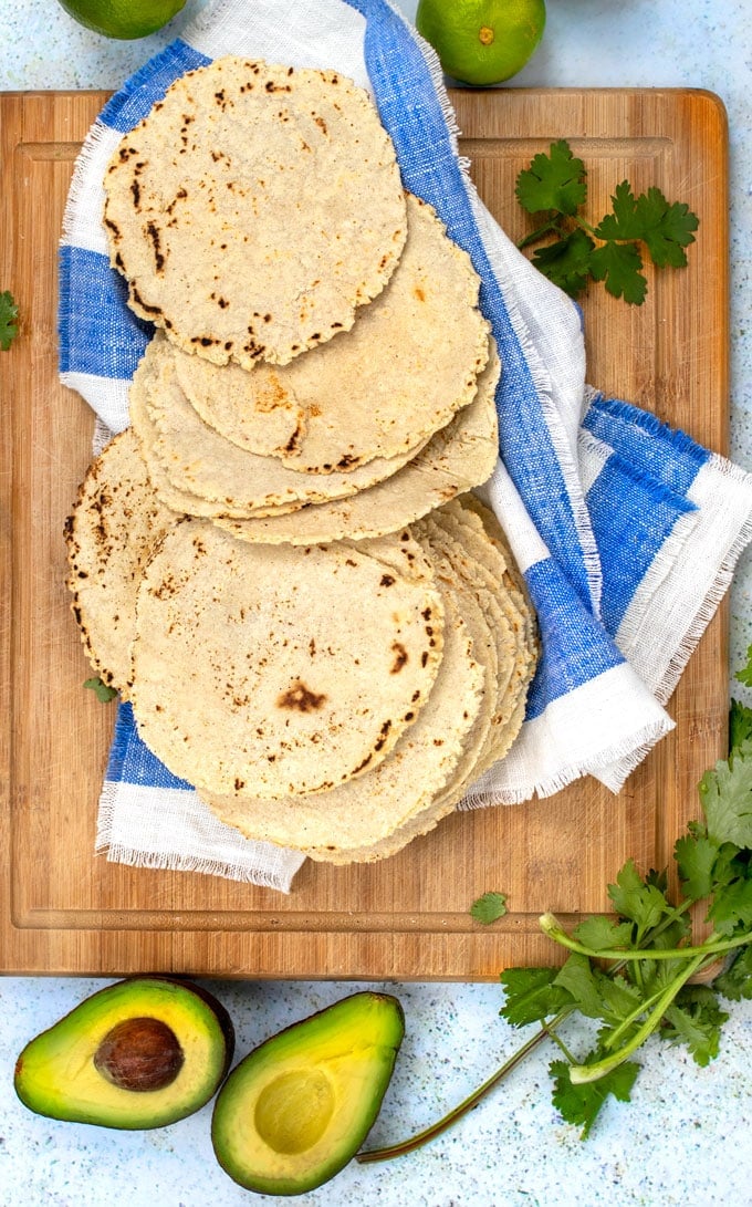 homemade corn tortillas stacked and spread out on a blue and white striped dish towel with some cilantro scattered around and a halved avocado