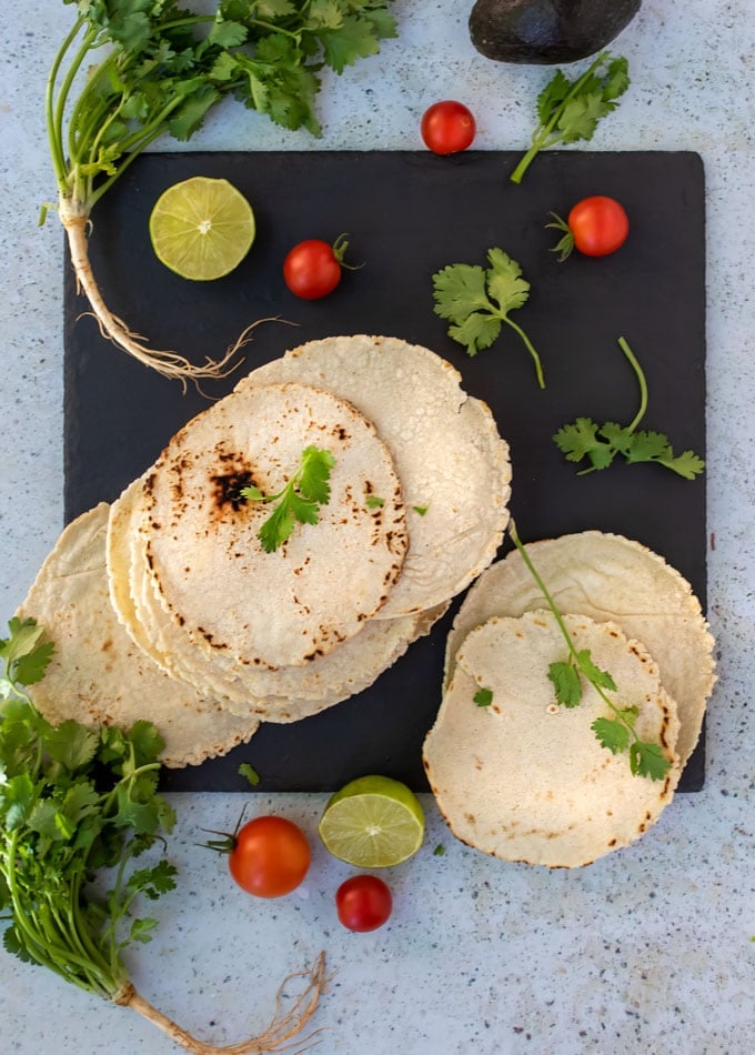 a stack on homemade corn tortillas on a black slate board, surrounded by sprigs of cilantro, cherry tomatoes and two lime halves