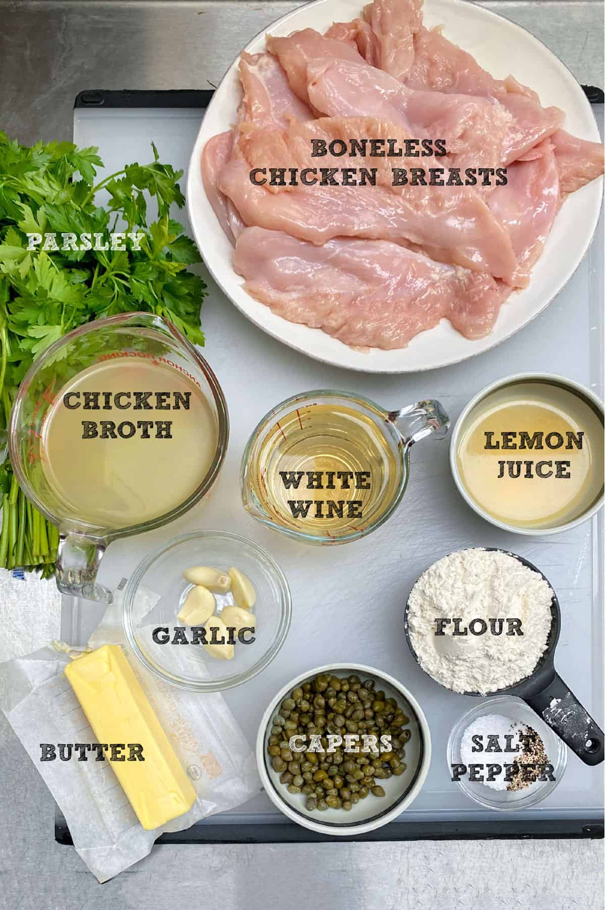 Ingredients for chicken piccata seen from above in small bowls and plates: pounded chicken cutlets, a bunch of parsley, white wine, lemon juice, chicken stock, a few cloves a garlic, a bar of butter, white flour, capers, salt and pepper