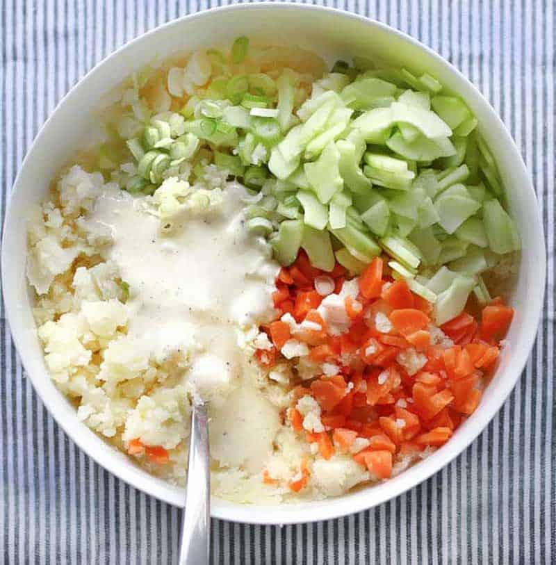 white bowl with ingredients for Japanese potato salad: mashed potatoes, chopped carrots, sliced cucumbers, scallions and mayo dressing