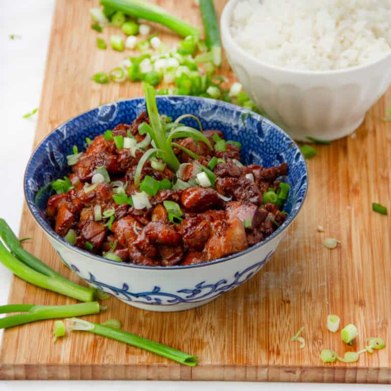 Asian honey soy chicken topped with sliced scallions, in a blue china bowl with a bowl of white rice in the background.