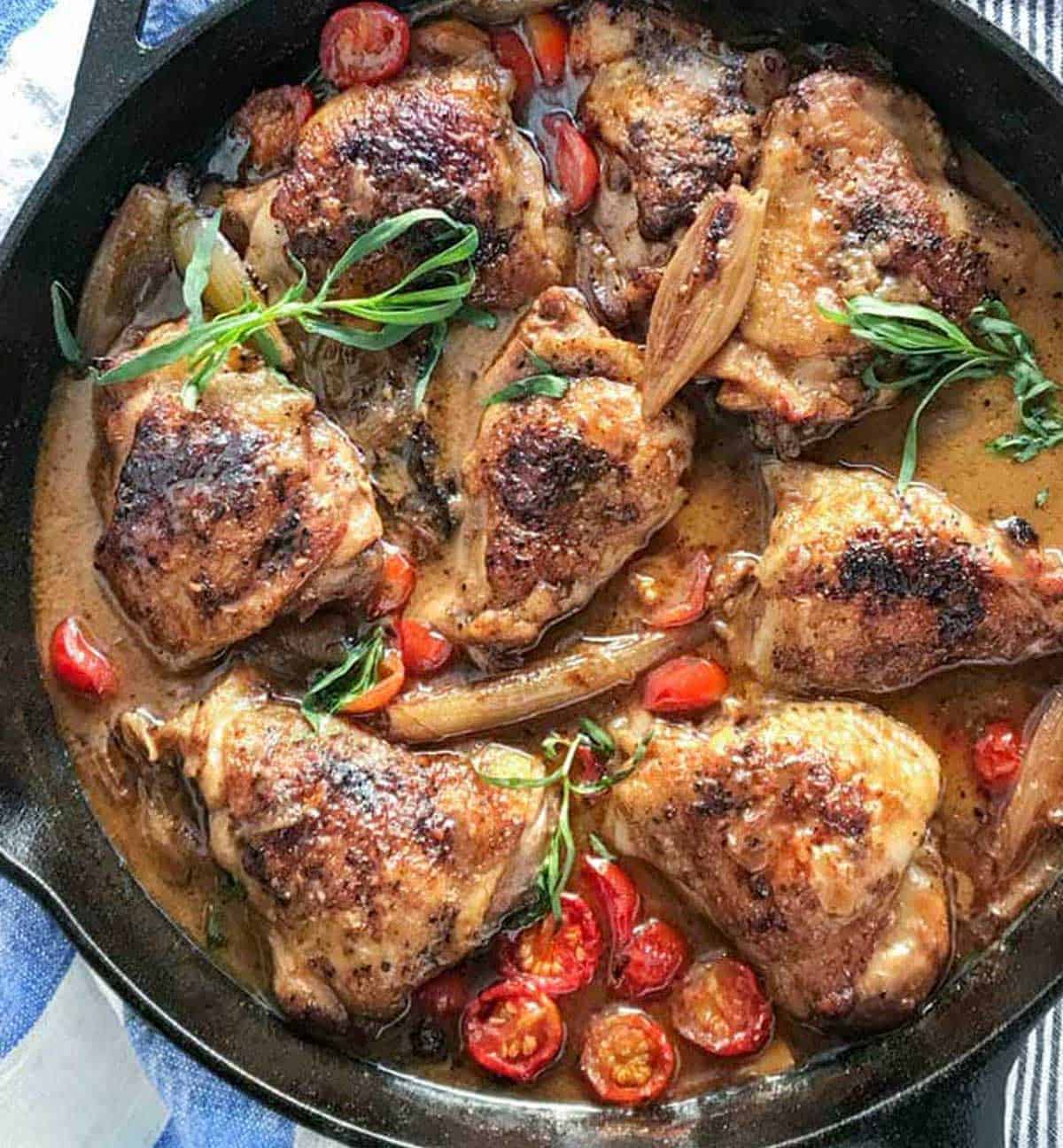 5 braised chicken thighs in a cast iron skillet with cherry tomatoes, shallots and sprigs of tarragon