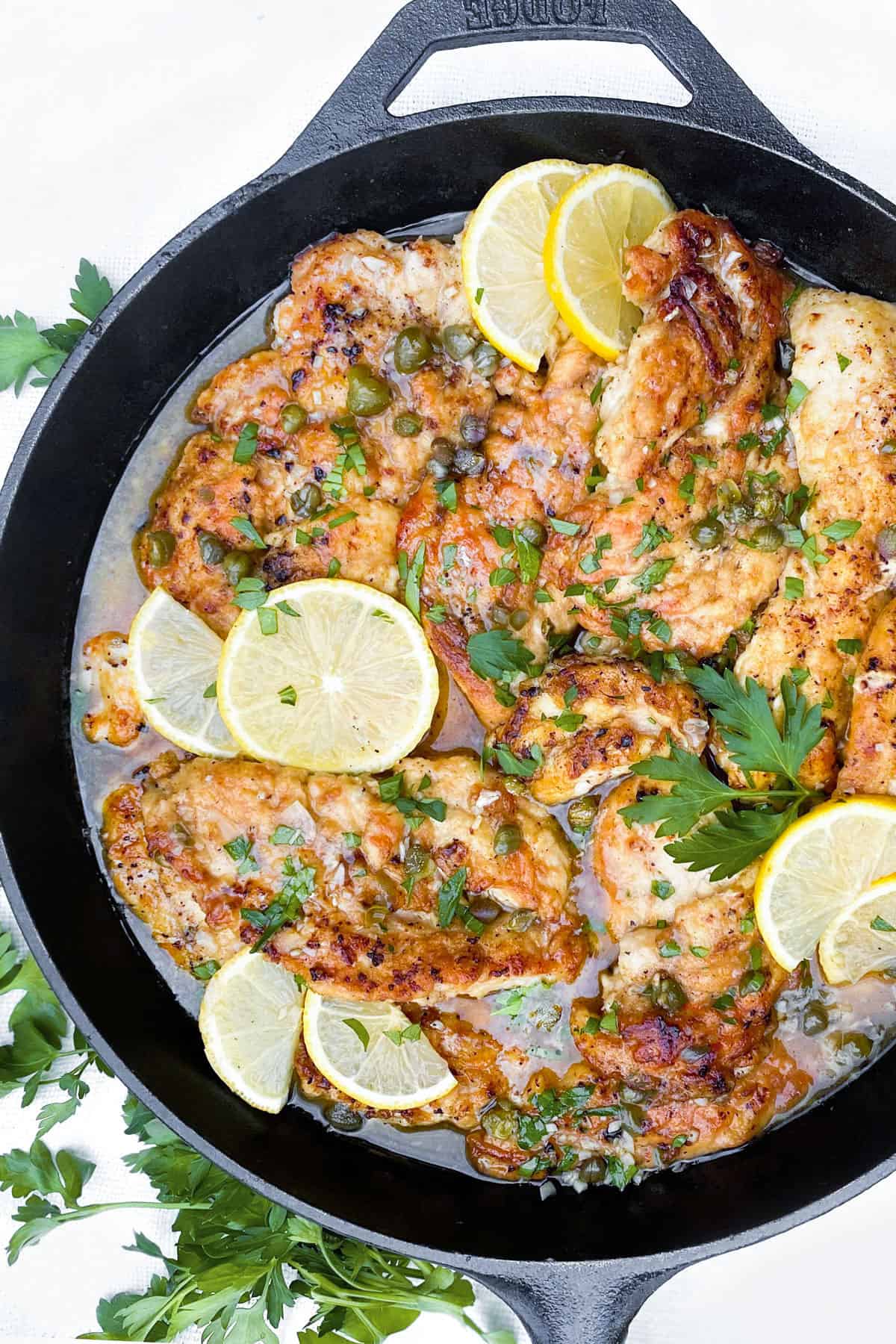 overhead shot of a black cast iron skillet filled with chicken piccata, thin chicken cutlets with a few thin slices of lemon tossed on top and some chopped parsley