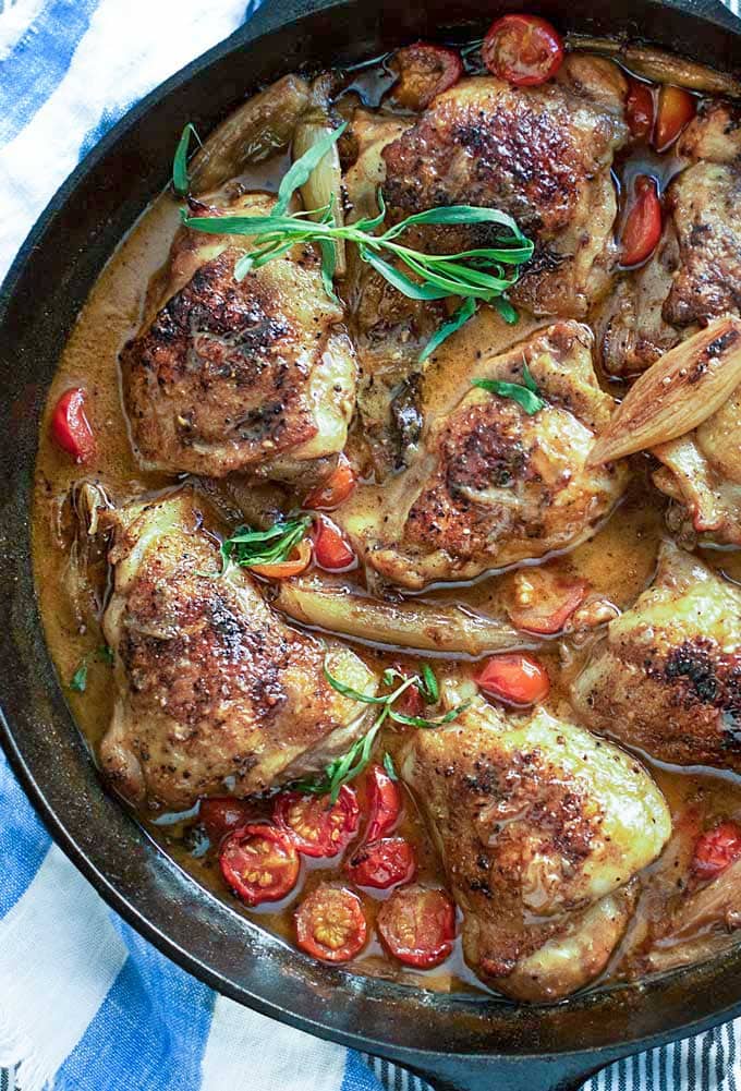 cast iron skillet with 8 braised chicken thighs in mustard wine sauce with sliced cherry tomatoes and sprigs of tarragon