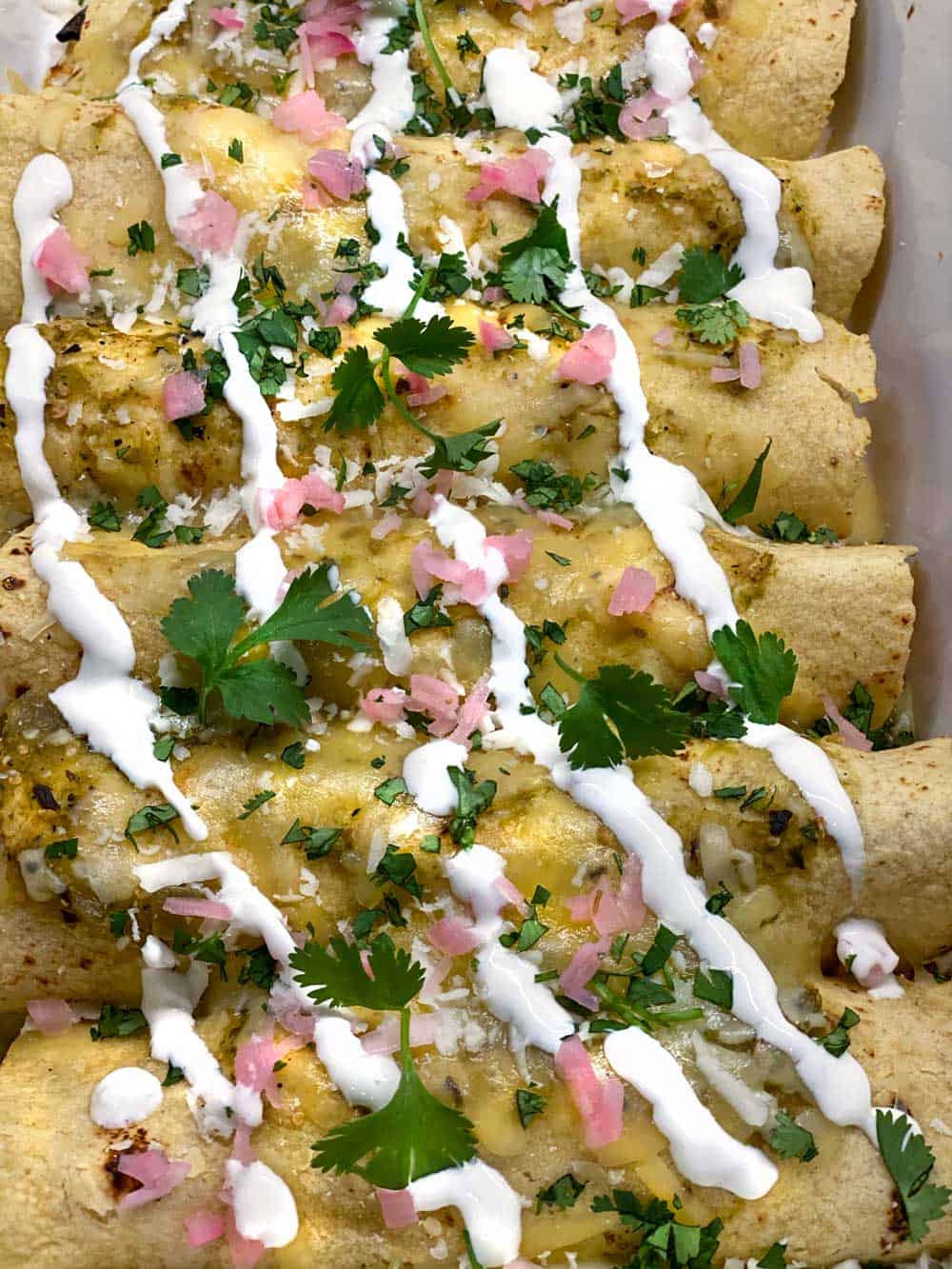 Close up of 6 vegetarian enchiladas verdes topped with stripes of sour cream, sprinkles of chopped cilantro and chopped pickled red onions.