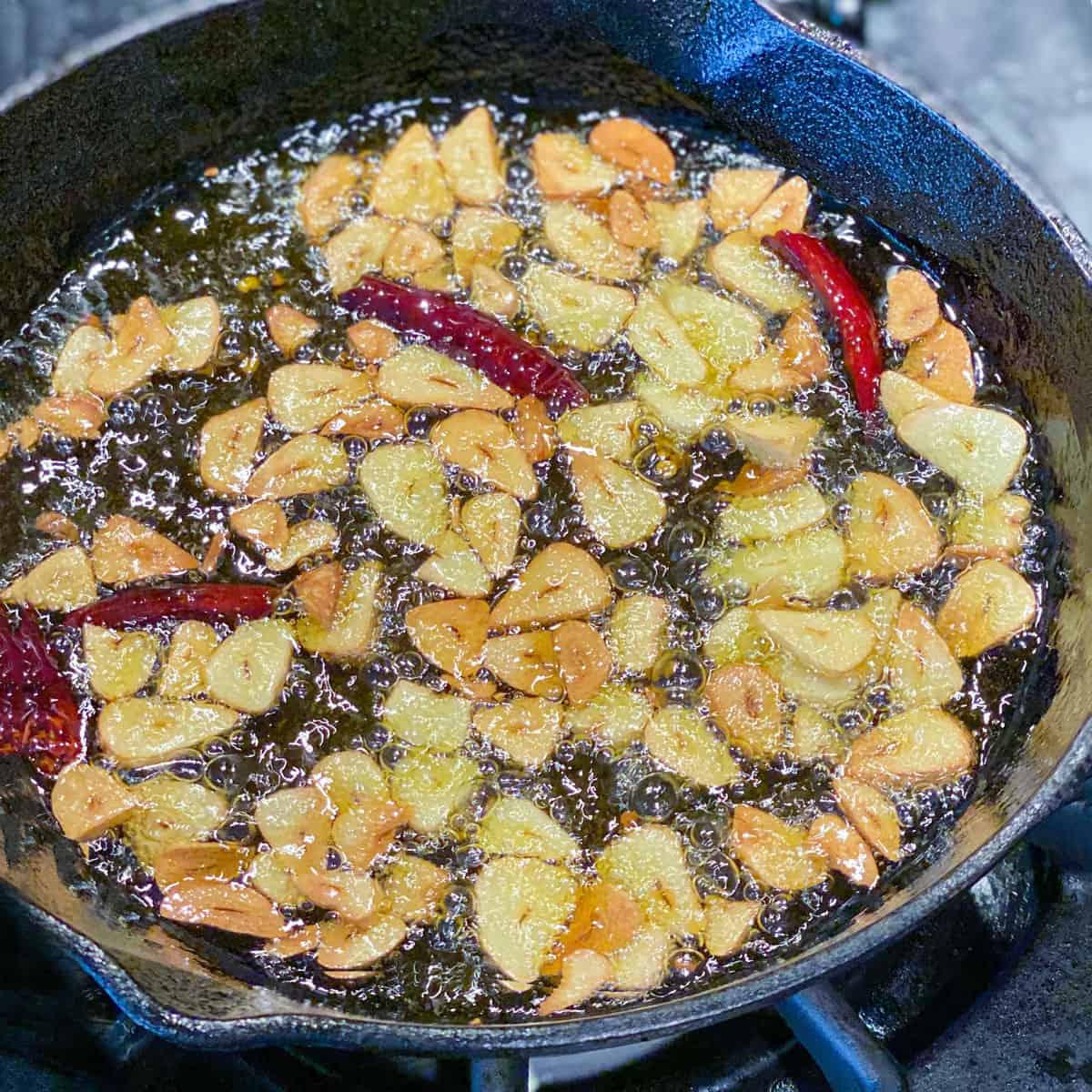 Golden brown slices of garlic in olive oil in a small cast iron skillet, with 3 small red chili peppers.