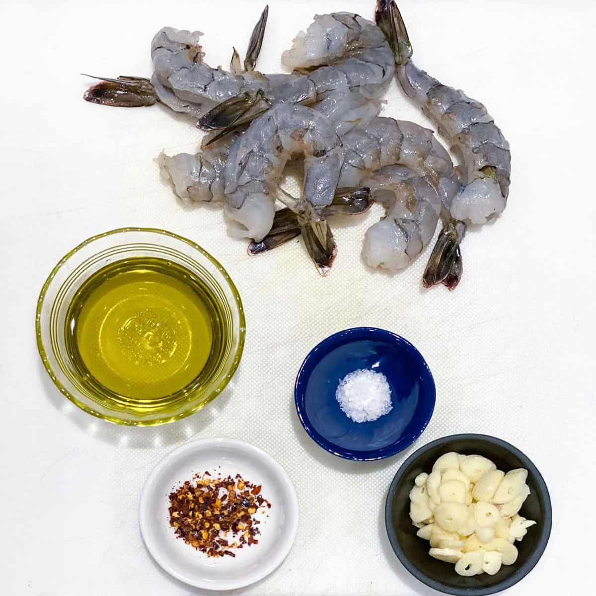 Ingredients on a white countertop: a handful of raw peeled shrimp, a small bowl of olive oil, small bowl of chopped garlic, salt in a small bowl, red pepper flakes in a small bowl.