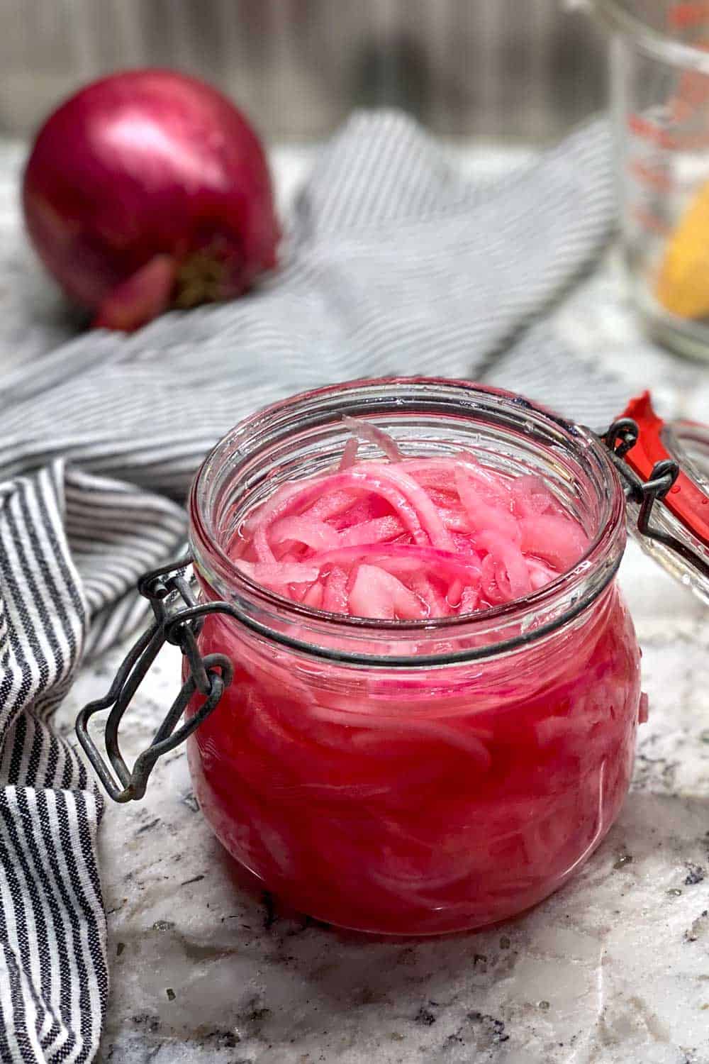 glass jar of filled with pickled red onions sitting on a white marble countertop with a striped dish towel and a whole red onion in the background