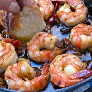 8 sauteed shirmp in a small cast iron skillet with olive oil, garlic and red chili pepper, a hand dipping a piece of sliced baguette into the oil.