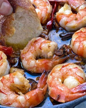 8 sauteed shirmp in a small cast iron skillet with olive oil, garlic and red chili pepper, a hand dipping a piece of sliced baguette into the oil.