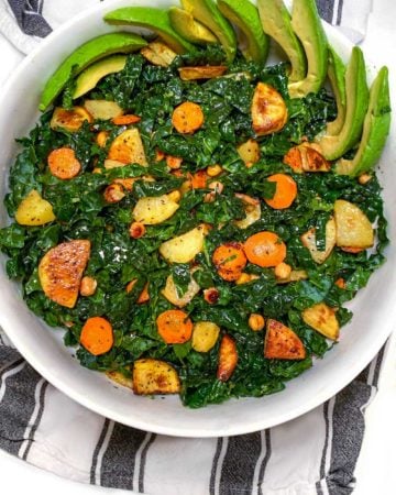white serving bowl on black and white striped dish towel, filled with shredded kale and roasted carrots, potatoes, chickpeas and sliced avocado