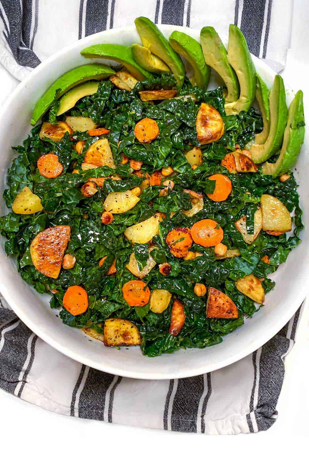 white serving bowl on black and white striped dish towel, filled with shredded kale and roasted carrots, potatoes, chickpeas and sliced avocado