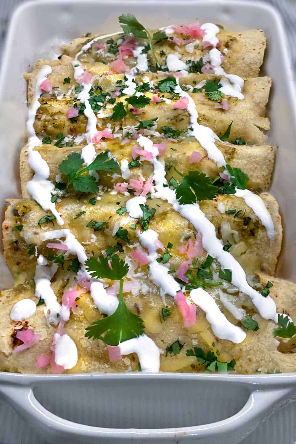 a white baking pan filled with a row of 6 vegetarian enchiladas verdes topped with stripes of sour cream, sprinkles of chopped cilantro and chopped pickled red onions.