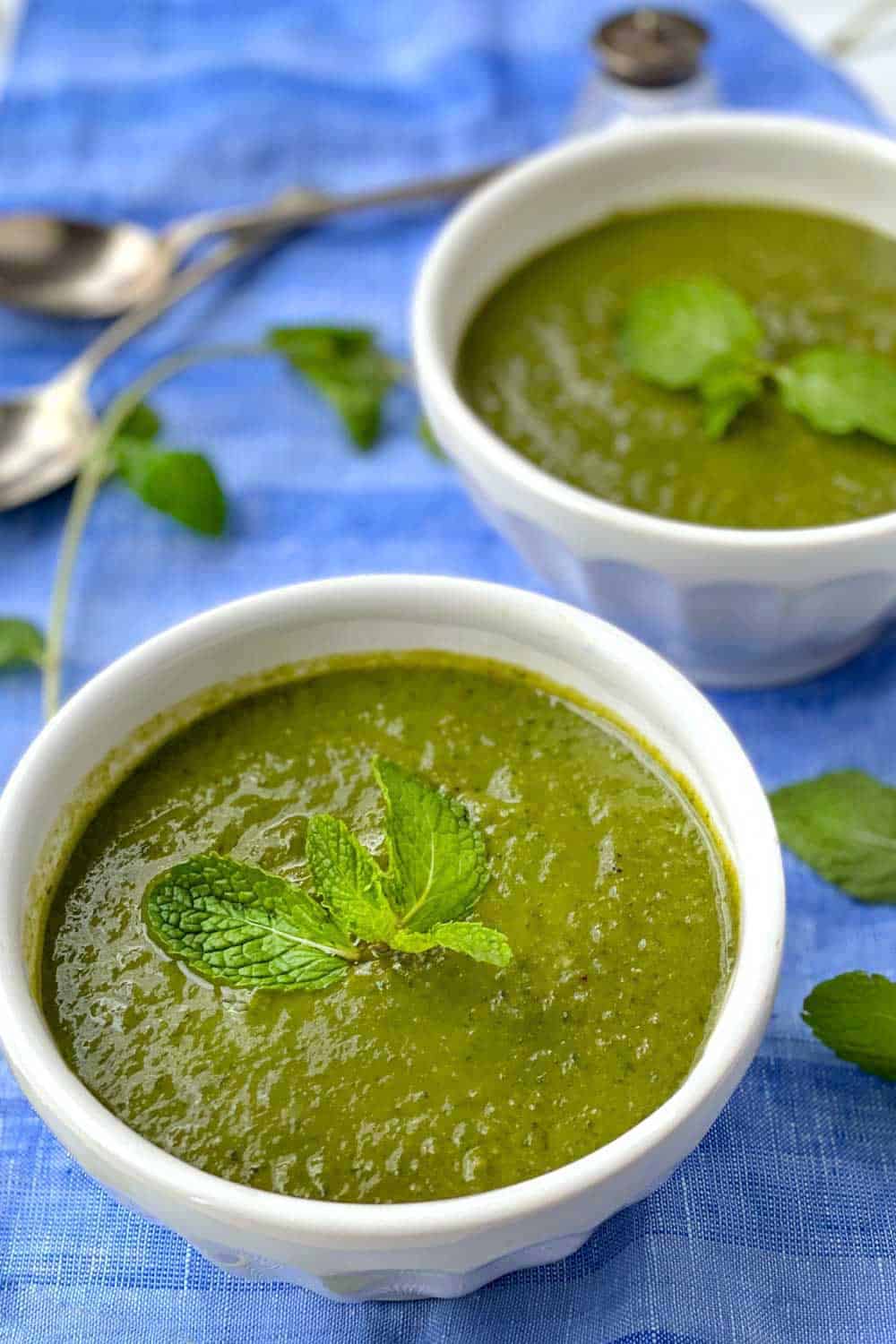 two white bowls filled with minted pea and spinach soup, sitting on a vivid blue table cloth