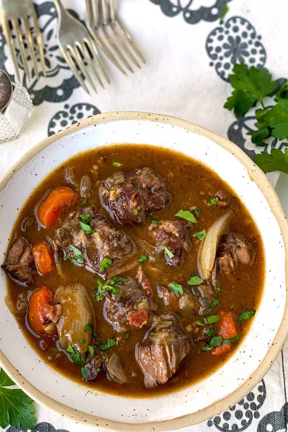 A white bowl filled with beef bourguignon sits on a black and shite flowered dish towel with four forks in the background and a sprig of parsley