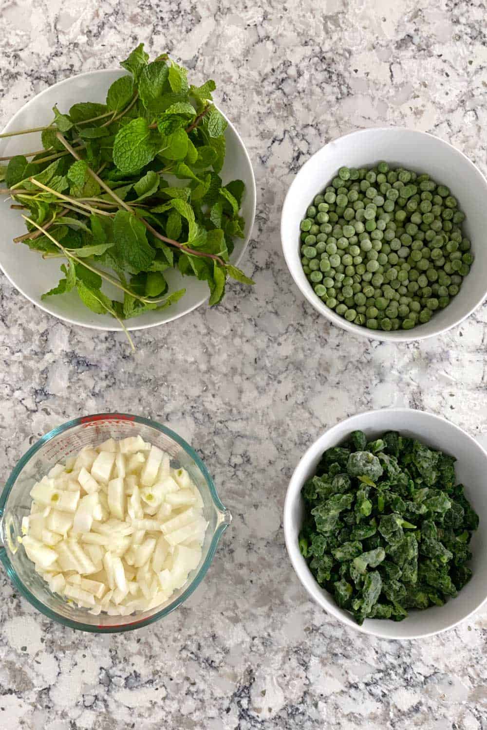 ingredients for pea and spinach soup with fresh mint: a bowl of frozen peas, a bowl of frozen spinach, a bowl of fresh mint sprigs and a measuring cup with chopped onions.