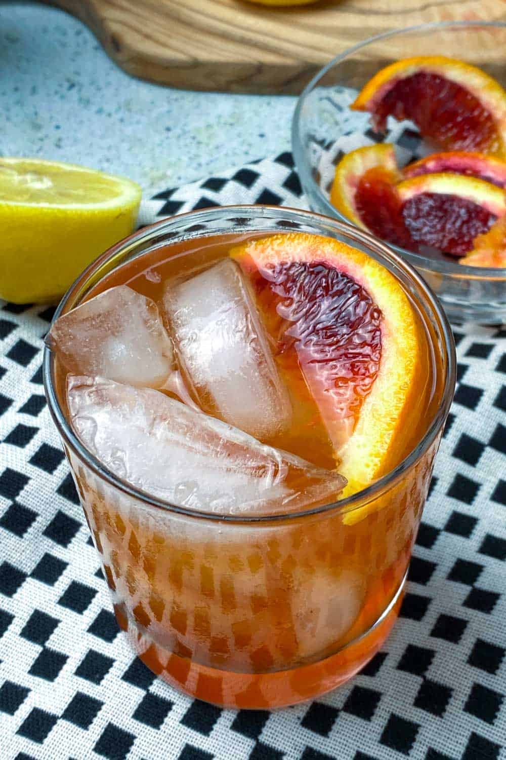 overhead view of a paper plane cocktail with ice cubes and a slice of blood orange. The glass sits on a vivid white dishtowel with black squares