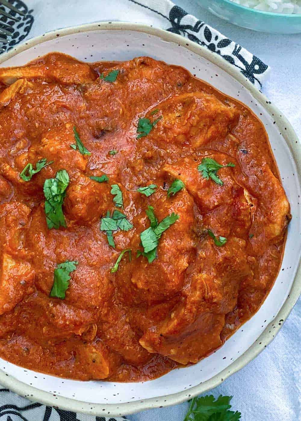 close up of a white bowl filled with vivid tomato-based chicken ruby, Indian chicken curry, with a sprinkle of parsley on top